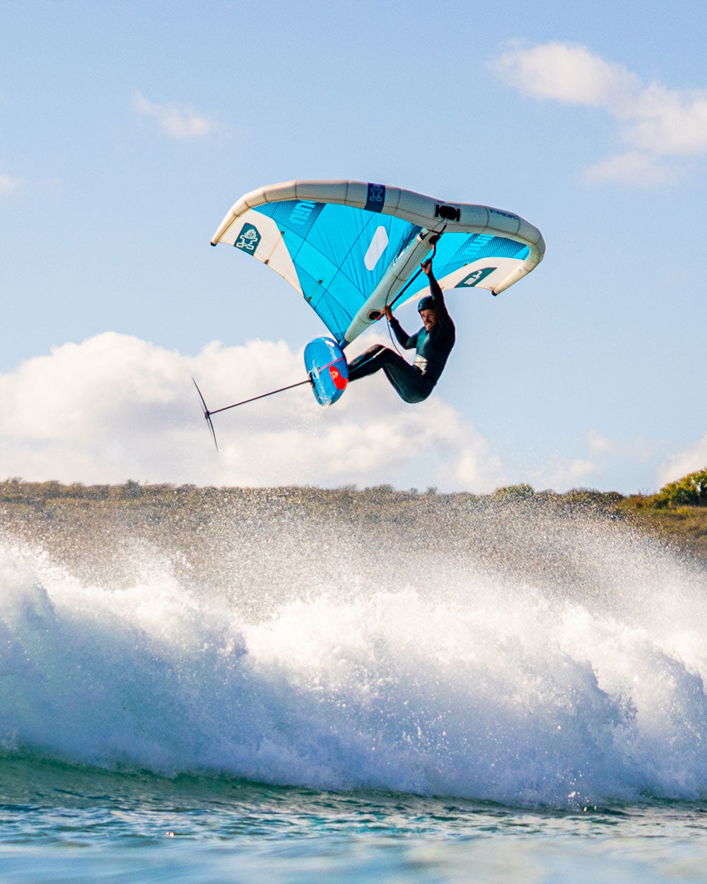 Viva Wing rider in black wetsuit jumps mid-air on blue and white sail above crashing waves