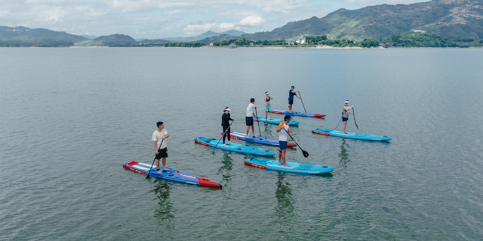 Group of seven paddlers standing on Starboard inflatable paddle boards on calm water with green mountains in the background