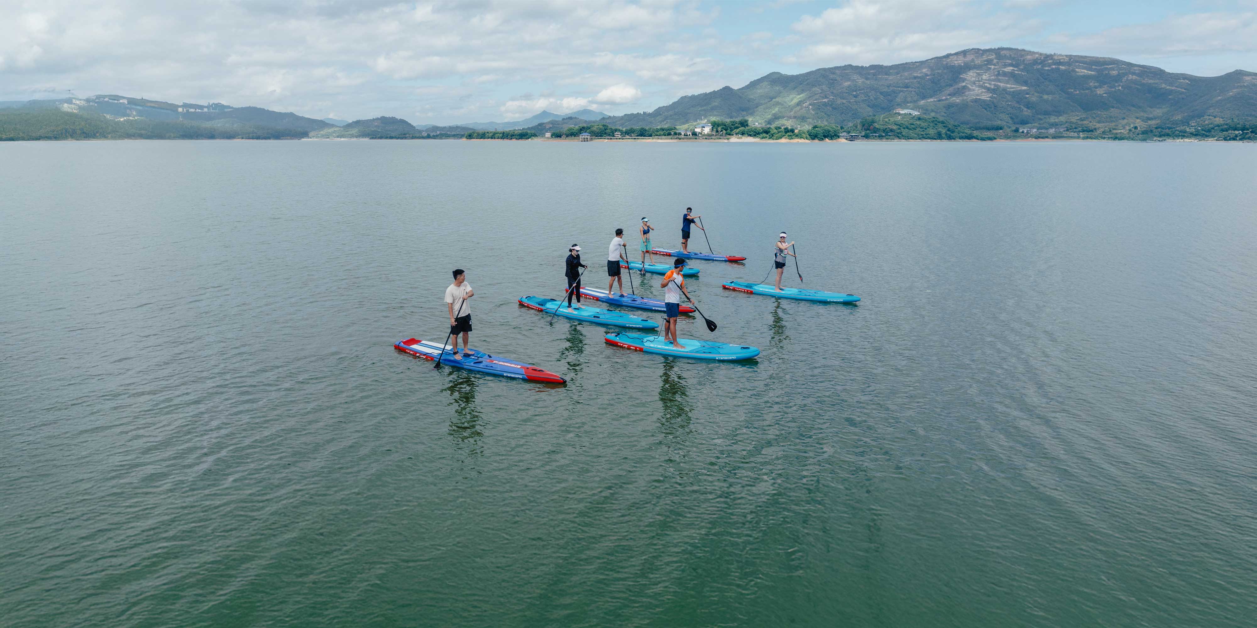Group of seven paddlers standing on Starboard inflatable paddle boards on calm water with green mountains in the background